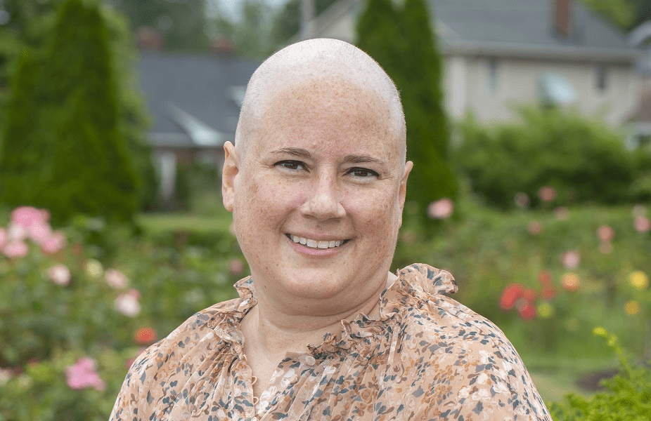 A bald, smiling woman sits outside on a park bench during a summer day.