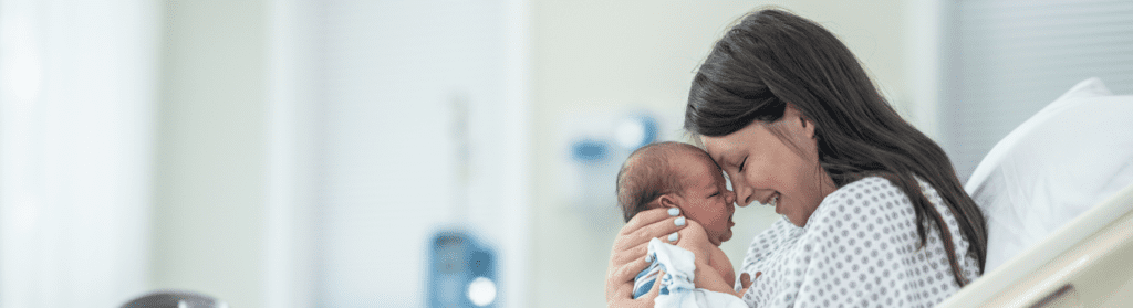 A young woman smiles while holding her newborn infant to her in a hospital bed.