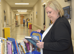 A middle-aged woman browses a selection of books on a blue cart in a hospital room.