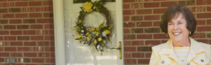 An older woman in a yellow jacket stands outside a red brick house.