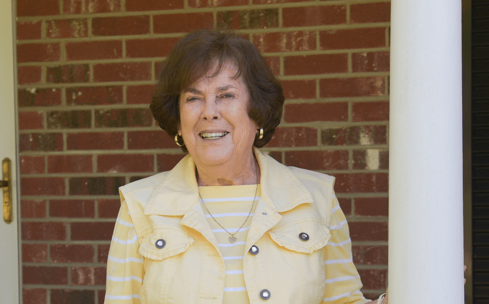 An older woman in a yellow jacket stands outside a red brick house.