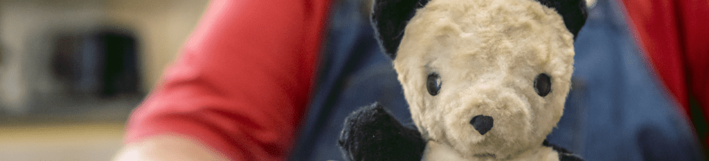 An older man in overalls sits inside at his kitchen table, holding an aged stuffed teddy bear.