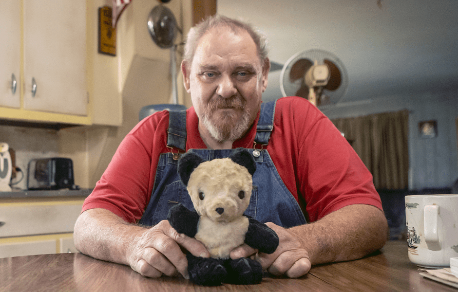 An older man in overalls sits inside at his kitchen table, holding an aged stuffed teddy bear.
