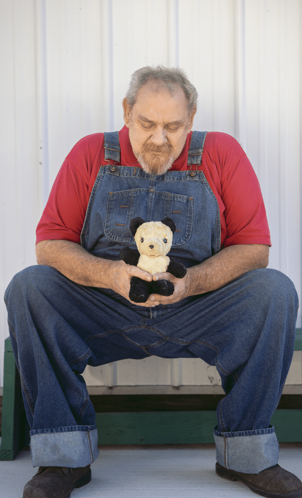 An older man in overalls sits outside on a stoop, holding an aged stuffed teddy bear.
