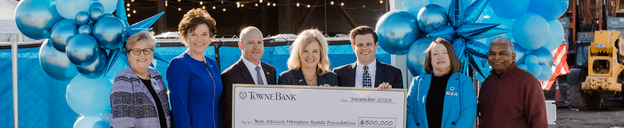 A group of business people, surrounded by balloons, exchange an XL check in front of a hospital under construction.