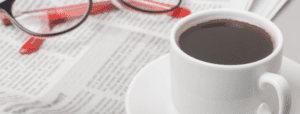 A cup of coffee and a pair of glasses rest atop an unfolded newspaper.