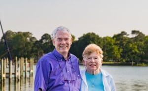 An elderly couple stands together beside a lake on a sunny day.