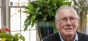 An elderly man with glasses sits inside among some house plants.