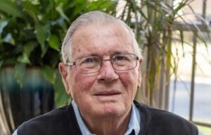 An elderly man with glasses sits inside among some house plants.
