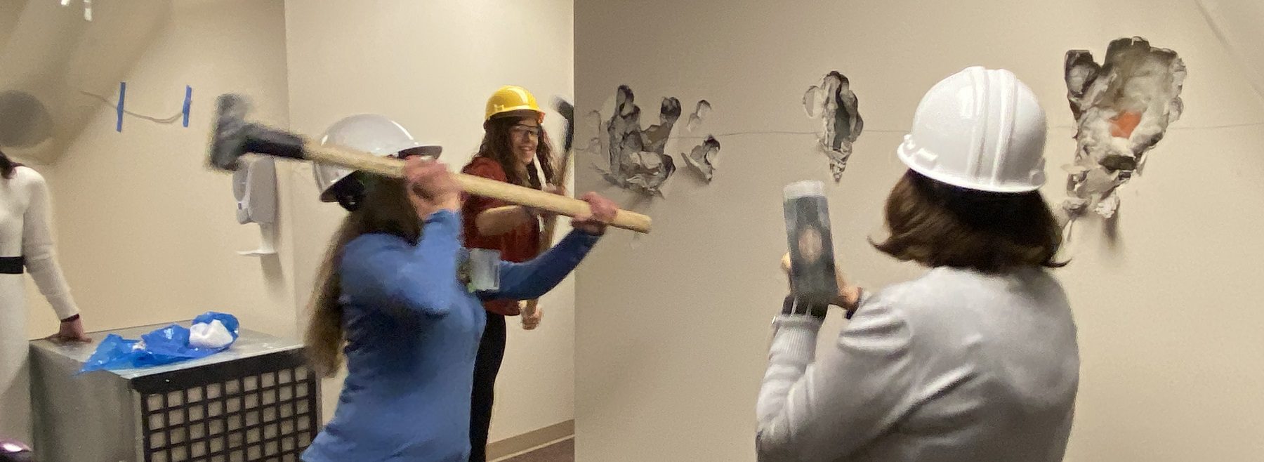 Professional women wearing hard hats swing sledgehammers at a wall.