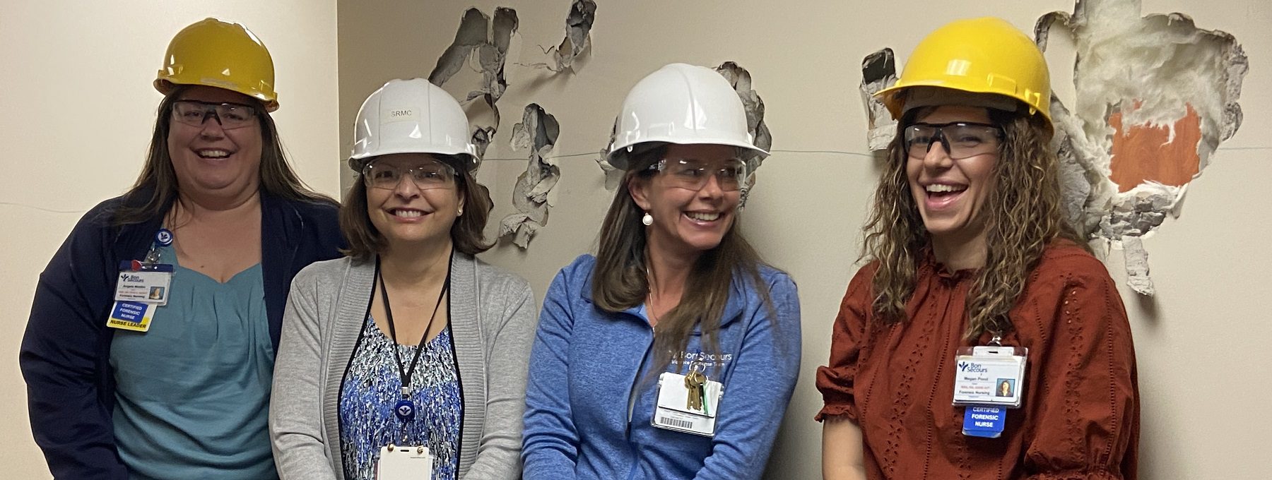 A group of professional women wearing hard hats pose and smile underneath a row of holes in drywall.