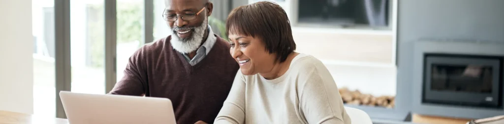 A middle-aged couple sits at a table together, working at a laptop.