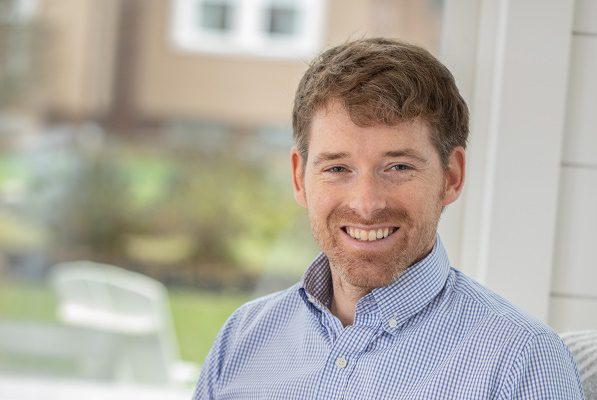 A young businessman sits inside his home, smiling.