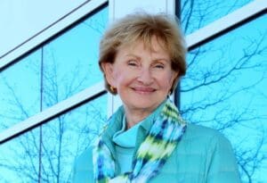 An elderly woman in a bright blue sweater stands outside, smiling.