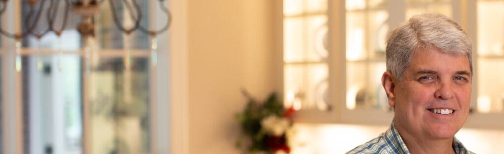 An elderly man stands in his sunny dining room, smiling.