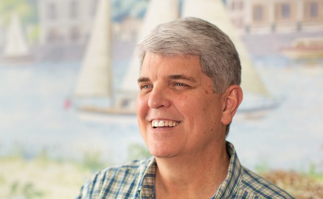 An elderly man sits in front of an indoor mural that depicts sailboats.