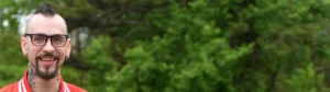 A man with a mohawk, glasses, and goatee stands outside in a park, smiling.