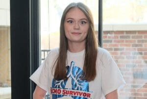 A teenage girl sits inside a medical facility. She is wearing a t-shirt that reads "ECMO Survivor."