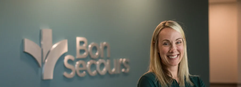 A smiling woman stands in front of a dark green Bon Secours sign.