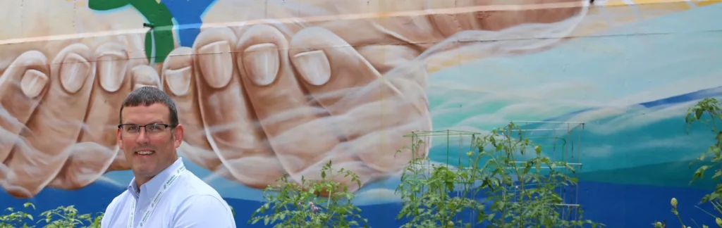 A man in a dress shirt sits in front of a mural of the hands of Jesus. The Bon Secours Mercy Health logo is cupped in the hands.