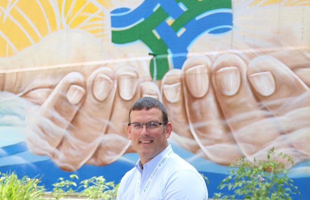 A man in a dress shirt sits in front of a mural of the hands of Jesus. The Bon Secours Mercy Health logo is cupped in the hands.