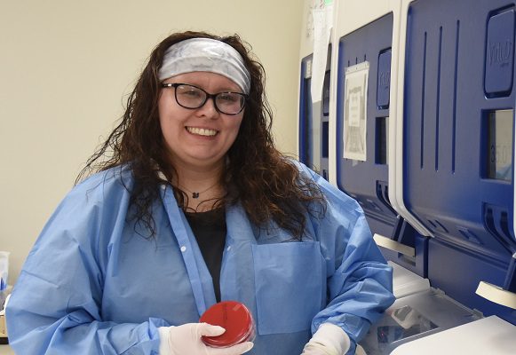 A young lab technician stands in front of a big lab machine. She's holding a culture sample in a petri dish.