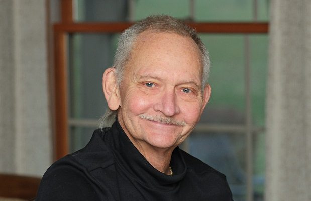 An elderly man in a black turtleneck stands in front of a window in his home.