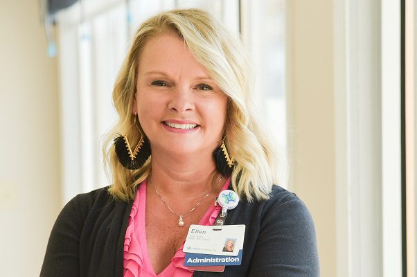 A middle-aged hospital administrator smiles in front of sunny windows.