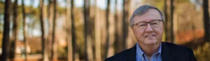 An elderly man stands outside in the woods on a sunny day, smiling.