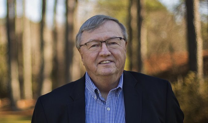 An elderly man stands outside in the woods on a sunny day, smiling.