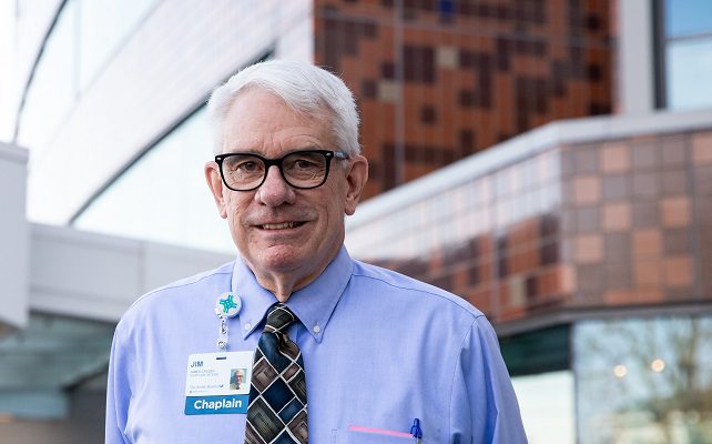 An older man with a Chaplain tag on his shirt stands outside Mercy Health - Jewish Hospital.