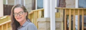 A middle-aged woman with long brown hair stands on her front porch on a sunny day.