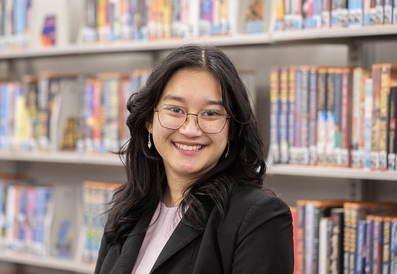 A young female student stands in front of bookshelves in a library.