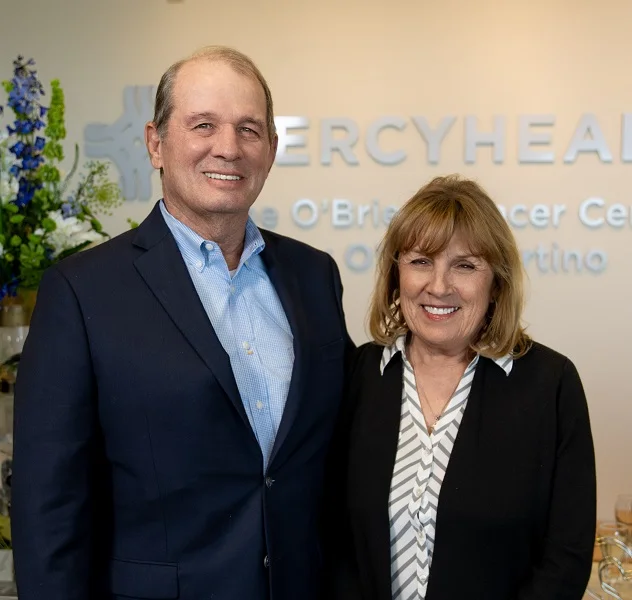 A middle-aged couple dressed in formal attire pose in front of a sign for Mercy Health – O’Brien Cancer Center.