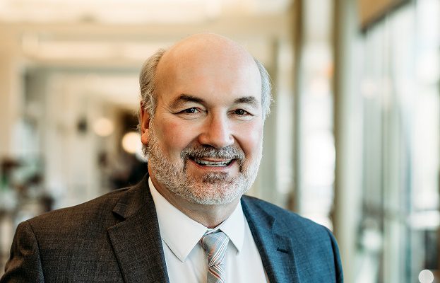 A bald man with a beard sits inside a hospital lounge. He's wearing a suit,