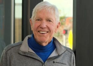 A senior man in a Mercy Health zip-up jacket smiles while standing in front of a window.