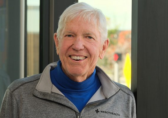 A senior man in a Mercy Health zip-up jacket smiles while standing in front of a window.