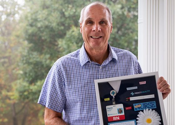 A middle-aged man stands on his porch, holding a shadow box of nursing memorabilia.