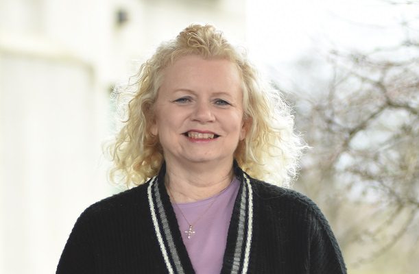 A smiling middle-aged woman stands outside on a sunny day.