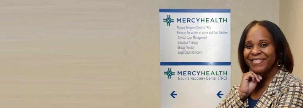A professional woman standing next to a sign for Mercy Health – Toledo Trauma Recovery Center.