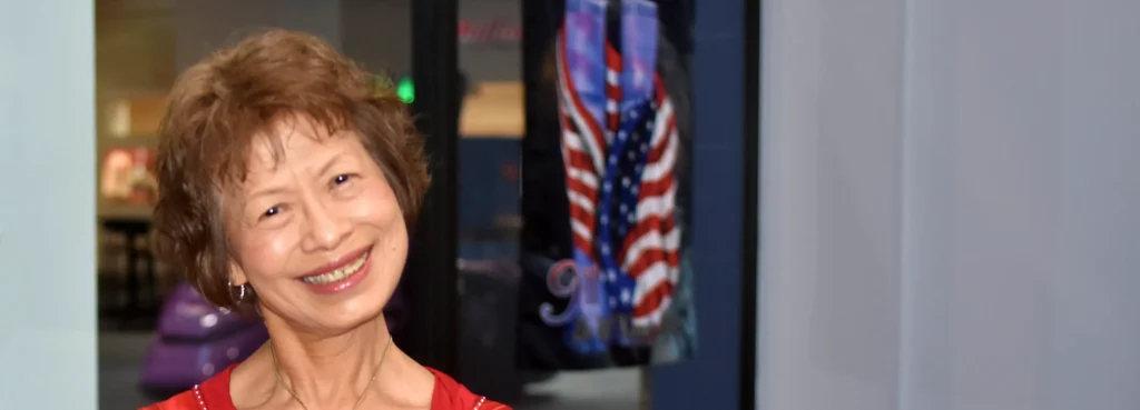 An elderly woman stands inside a gym, smiling at the camera with her head cocked happily.