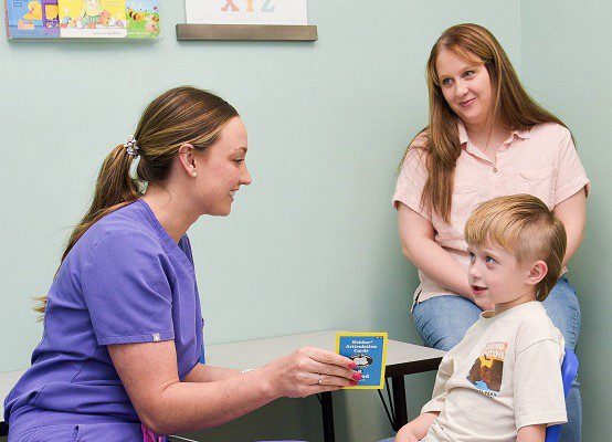 A speech therapist in purple scrubs does flashcards with a shy five-year-old boy.