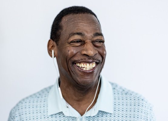 A man with a wide, bright smile listens to something with a pair of in-ear headphones.