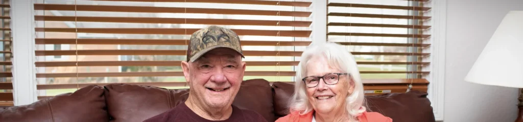 A senior couple sit on a brown leather sofa in their home.