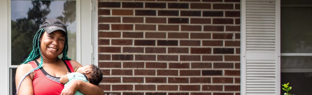 A young, new mom holds her infant in her arms while standing outside their brick house.