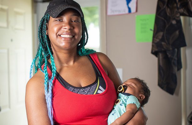 A young, new mom holds her infant in her arms while standing inside their house.