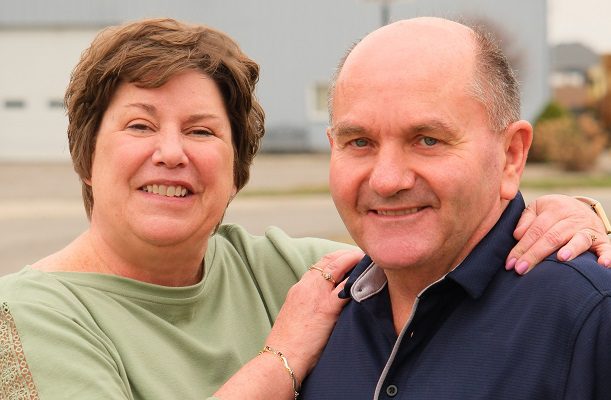 A middle aged couple stands outside on a sunny day, smiling. The woman has her arm around the man.