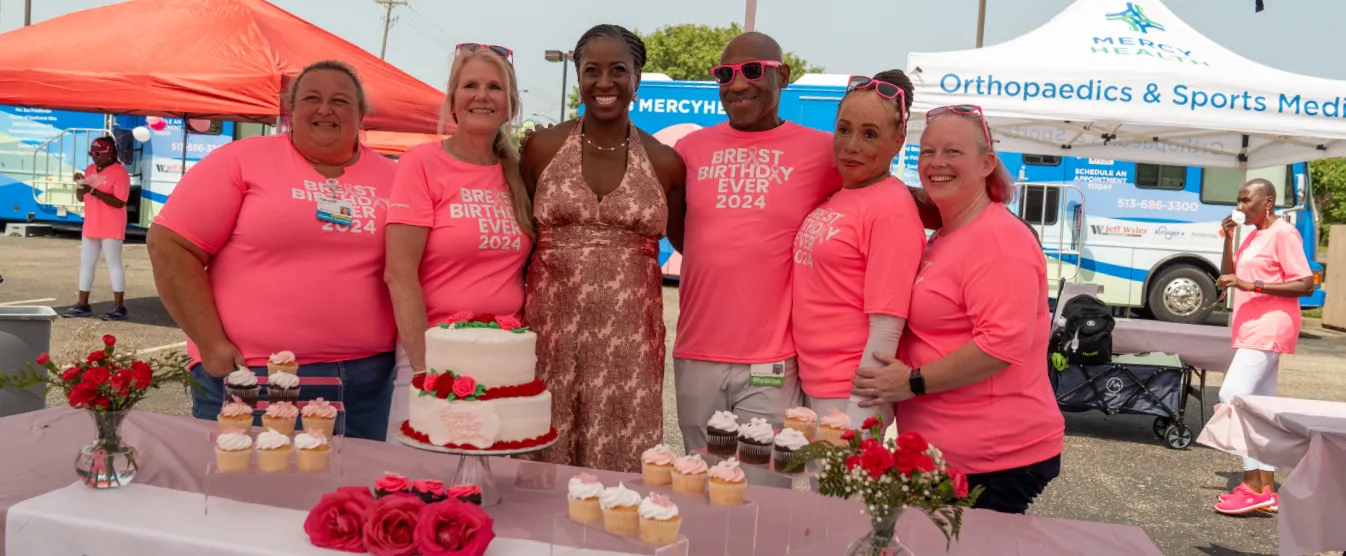 A group of people wearing bright pink shirts gather around a table full of cake and cupcakes.