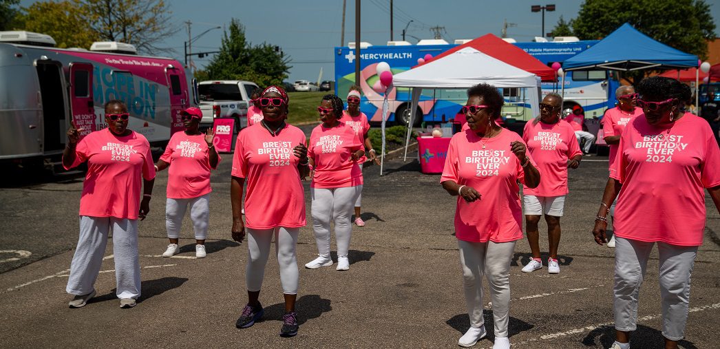 A group of women in bright pink shirts dance together outside on a sunny day.