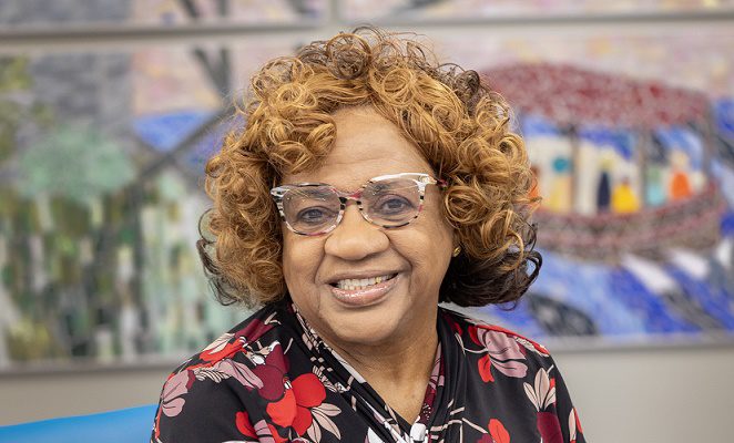 An elderly woman in a floral blouse smiles while sitting in front of a mural.